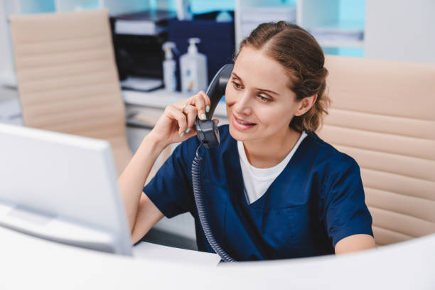 unga kvinnliga receptionist talar i telefon på kliniken medan du sitter och tittar på pc monitor - happy scrubs nurse phone bildbanksfoton och bilder