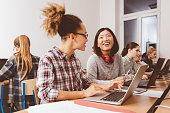 Multiracial group of female students coding on laptops in a computer lab. Asian girl talking to her afro american friend and smiling.