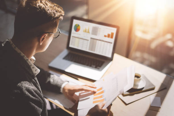 Young finance market analyst in eyeglasses working at sunny office on laptop while sitting at wooden table.Businessman analyze document in his hands.Graphs and diagramm on notebook screen.Blurred. stock photo