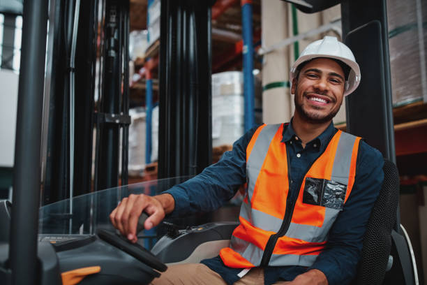 young forklift driver sitting in vehicle in warehouse smiling looking at camera - forklift imagens e fotografias de stock