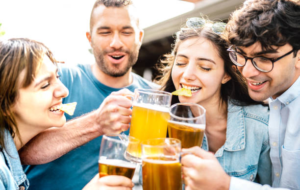 Young friends drinking and toasting beer pints at brewery bar garden out side - Beverage life style concept with men and women having fun together at happy hour - Bright filter with focus on left girl stock photo