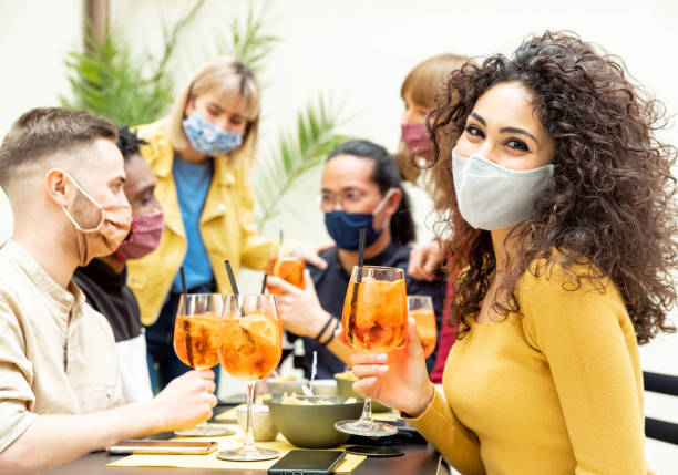 Young friends drinking cocktails wearing protective face mask - New normal lifestyle concept with people enjoying happy hour at cocktail bar - Food, drink and people concept. stock photo