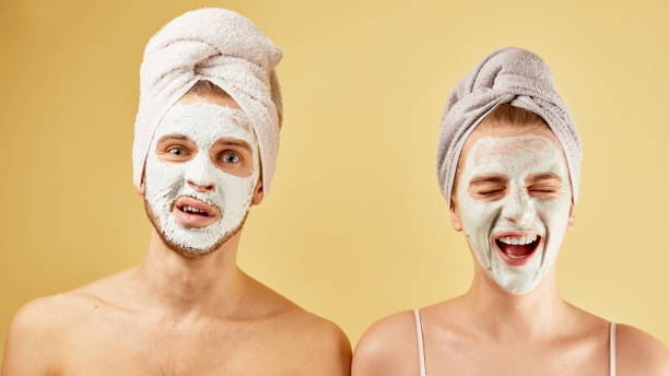 Young friends guy and girl with funny expressive emotions, towel on head and face mask isolated on a yellow background, skin care concept stock photo