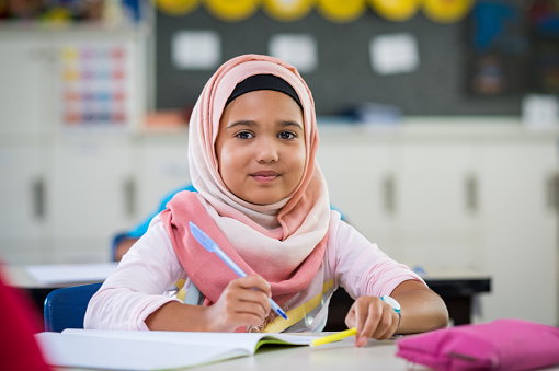 Happy young smiling girl wearing hijab and looking at camera while sitting at desk with book and pen in hand. Elementary muslim schoolgirl writing notes in classroom. Portrait of arab school girl in chador.