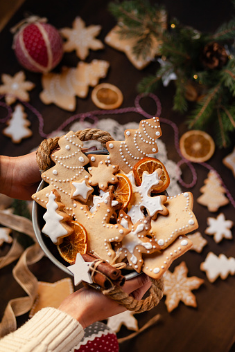 Young girl in knitted white sweater is holding a basket with homemade gingerbread cookies decorated with snow white icing. Anise and cinnamon as decor. Close up.