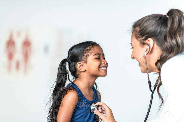 Young Girl is Examined by Female Doctor stock photo A young East Indian girl is examined by her Hispanic female doctor.  The doctor is checking her heart with the stethoscope that is around her neck.  The young girl is wearing a blue tank top while the doctor is wearing a white lab coat. pediatrician stock pictures, royalty-free photos & images