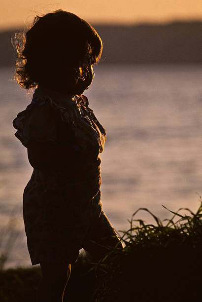 Young Girl Silhouetted at Sunset Puget Sound is a special body of water that belongs exclusively to Washington State. From the saltwater marshland of Olympia and Nisqually to the windy Strait of Juan de Fuca and the San Juan Islands, Puget Sound exhibits a quiet beauty that is quintessentially Washington. Puget Sound and its adjacent waters form a vast inland extension to the Pacific Ocean. Its complex network of straits, bays, canals and inlets give Western Washington a distinctive personality. Puget Sound along with its surrounding mountains has a major effect on the region's temperate climate. The area was first visited by Europeans in 1792 with British explorer George Vancouver's expedition. Captain Vancouver gave the region many of the place names which are still in use today. Not only does Puget Sound have a rich history, it also has some of the greatest scenery in the country. Sunsets over Puget Sound can be especially beautiful and an inspiration to photographers. This silhouette picture of a young girl was photographed as the sun set over Puget Sound from Saltwater State Park near Des Moines, Washington State, USA. jeff goulden sunset stock pictures, royalty-free photos & images