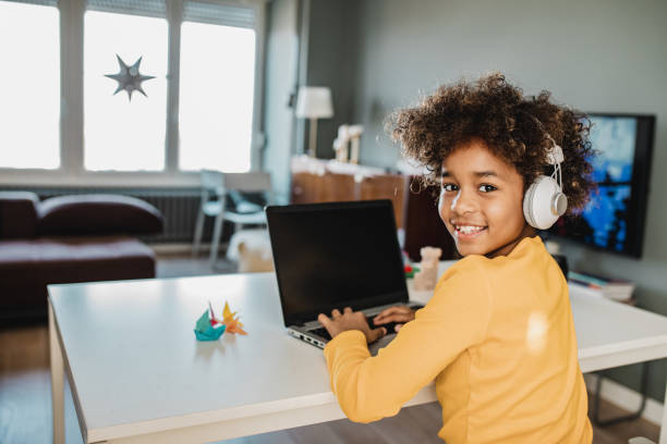 Young girl studying with laptop at home Young African american girl at home sitting on the table, using laptop, studying and looking at camera electrical equipment photos stock pictures, royalty-free photos & images
