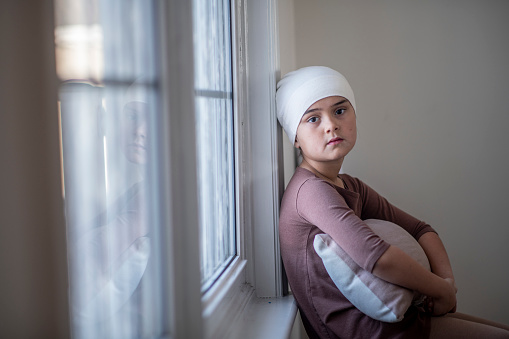 A young mixed race girl with cancer sits on a window ledge as she leans against it.  She is dressed casually and has a headcover on as she hugs a pillow and looks at the camera with a calm expression.
