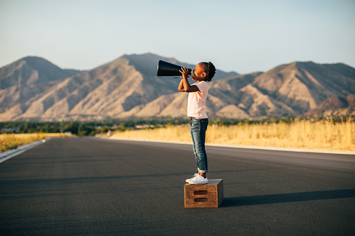 A young retro girl stands on a wooden box while yelling through a megaphone to have her message heard. Image taken in Utah, USA.