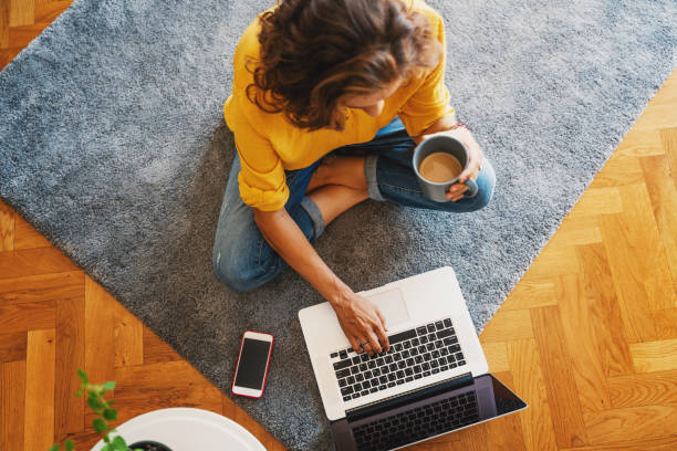 young girl woman in a yellow shirt works on a laptop with a mug of coffee at home in the living room on the carpet, remote work and education. Top view stock photo