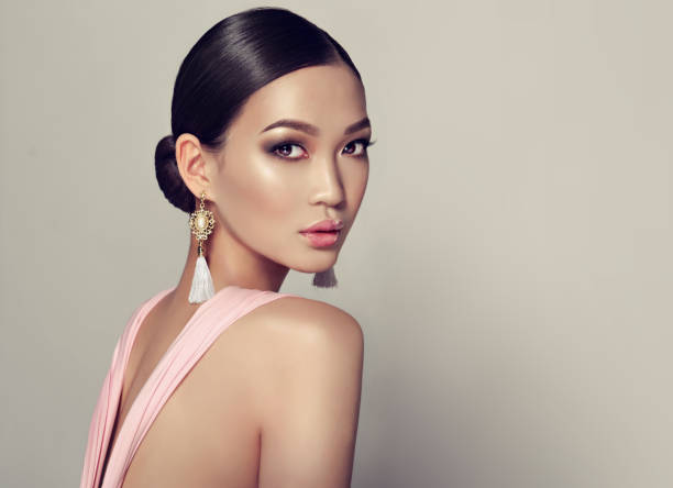 Young, gorgeous asian woman in a smoky eyes style make up, dressed in a tassel earrings. Young, gorgeous asian fashion-model put on in a smoky eyes style make up, black hair gattered in a beam. dressed in tassel earrings and  pink gown. Asian beauty. female likeness stock pictures, royalty-free photos & images