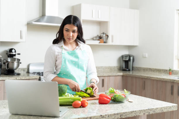 Young Gorgeous Woman Preparing Vegetable Salad In Kitchen Attractive housewife cutting bell pepper while learning through tutorial online from laptop at home cooking stock pictures, royalty-free photos & images