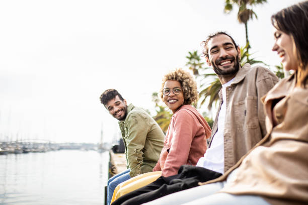 Young group of diverse adult friends laughing while sitting in front of the sea stock photo