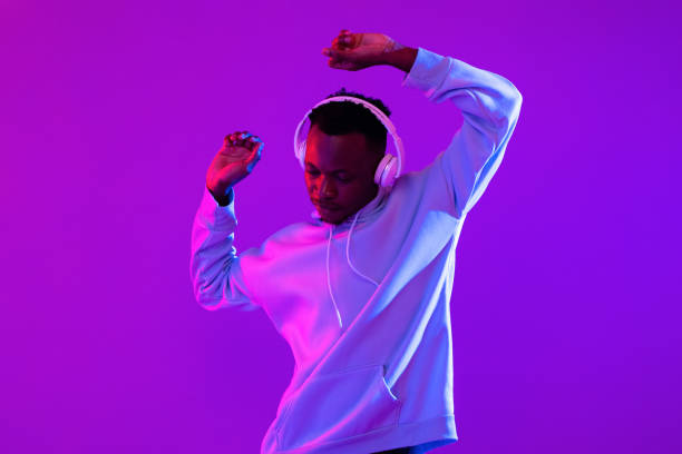 Young handsome African man wearing headphones listening to music and dancing in futuristic purple cyberpunk neon light background stock photo