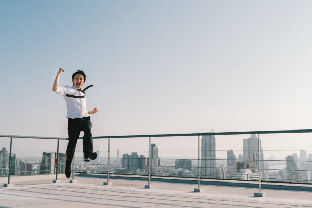 ジャンプ高い、若いハンサムなアジア系のビジネスマンは、建物の屋上に成功勝利ポーズを祝います。仕事、仕事、または巧妙なビジネス概念。晴れた青空にコピー スペースと都市景観の背 - ワクワク ストックフォトと画像