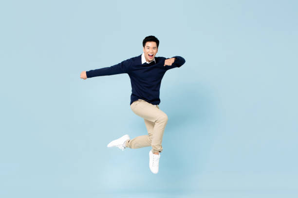 Young handsome Asian man smiling and jumping in mid-air on light blue studio background stock photo