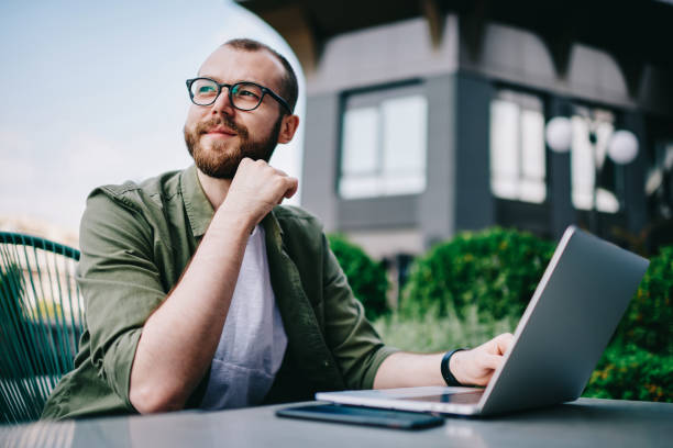Young handsome male 20 years old in eyeglasses sitting at street cafe during free time with new modern laptop, bearded man freelancer  thoughtfully looking aside and thinking about distance work stock photo