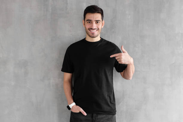 jonge knappe man die geïsoleerd op grijze geweven muur, lacht terwijl zwart t-shirt, copyspace voor reclame aan te wijzen met de wijsvinger - alleen één jonge man stockfoto's en -beelden