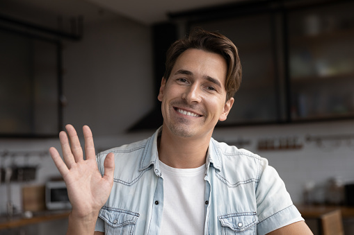 Head shot young handsome man waving hand looking at camera, starting online web call conversation. Confident happy millennial male blogger recording or streaming video, distant communication concept.