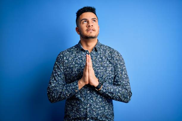 Young handsome man wearing casual shirt standing over isolated blue background begging and praying with hands together with hope expression on face very emotional and worried. Begging.  prayer request stock pictures, royalty-free photos & images