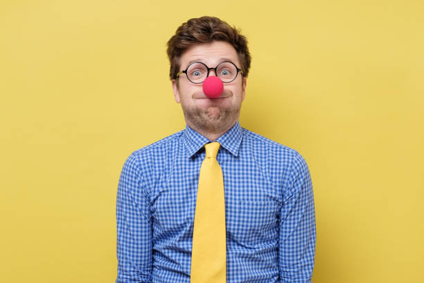 Young handsome man with red clown nose blowing cheeks. Young handsome man with red clown nose blowing cheeks. Studio shot on yellow wall clown's nose stock pictures, royalty-free photos & images