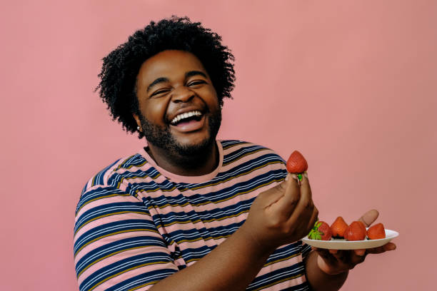 young happy african american man eating strawberries in the studio over pink background stock photo