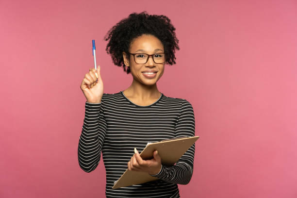 Young happy Afro-American teacher woman isolated on pink studio wall. Student girl wear glasses holding folder and pen, smiling, looking at camera. Education in high school university college stock photo
