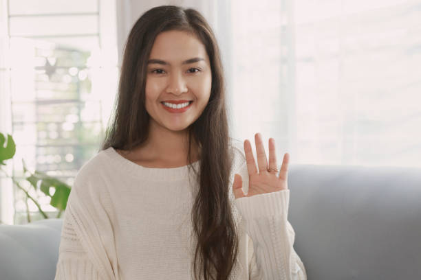 Young happy asian woman waving hand with smiley face while sitting on couch looking at camera. Asian woman greeting through the video call at home Young happy asian woman waving hand with smiley face while sitting on couch looking at camera. Asian woman greeting through the video call at home. wave goodbye asian stock pictures, royalty-free photos & images