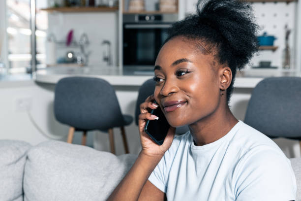 Young happy black African teenage woman sitting in her apartment talking on the smartphone hearing the good news from the company she applied for work over the internet application stock photo