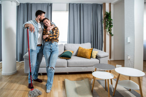 Young happy couple man and woman standing and smiling in their apartment where man hugs a woman and kiss her in her head after cleaning and rubbing floor of their home and finished housework together stock photo