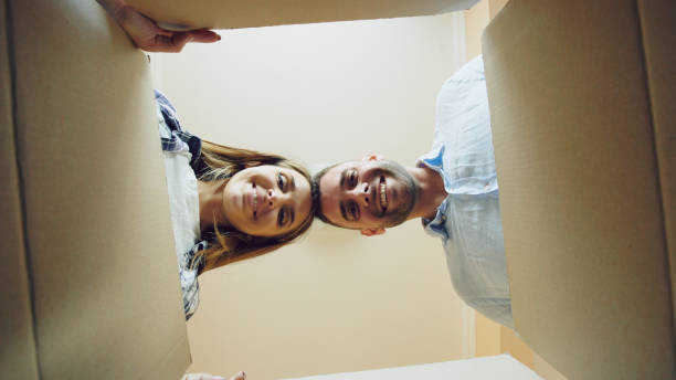 Young happy couple opening cardboard box after relocation and looking inside in new house stock photo