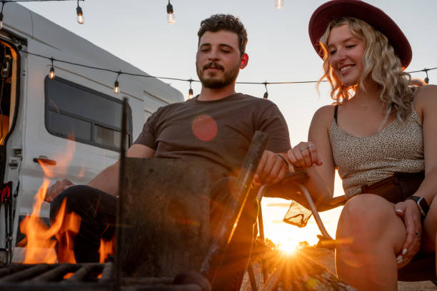 Young Happy Couple Sitting by a warm inviting campfire enjoying the Van life at a desert campsite at dusk stock photo