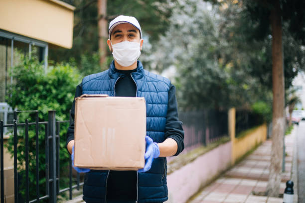 Young happy delivery man with protective mask looking at camera Young happy delivery man with protective mask looking at camera home delivery stock pictures, royalty-free photos & images