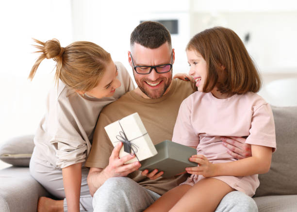 Young happy family celebrating holiday together at home Excited little daughter and her beautiful mother congratulating daddy with Fathers Day or birthday, giving him present wrapped gift box while sitting together on sofa at home fathers day stock pictures, royalty-free photos & images