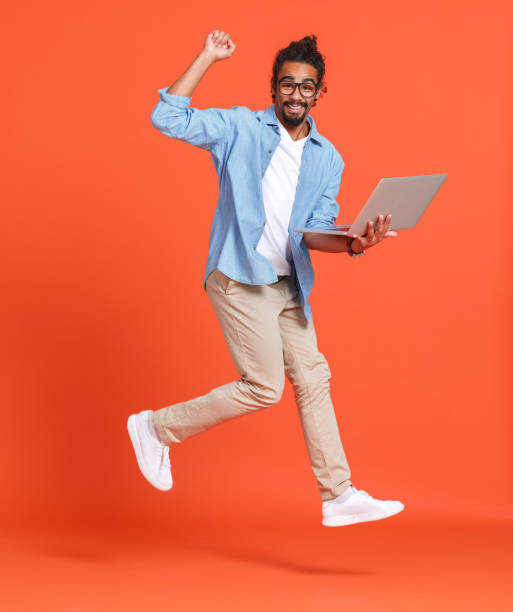 Young happy overjoyed african american man with laptop computer jumping over orange background stock photo