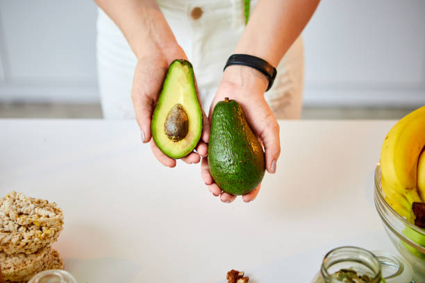 Young happy woman holding avocado for making salad in the beautiful kitchen with green fresh ingredients indoors. Healthy food and Dieting concept. Loosing Weight stock photo