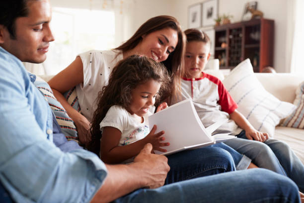 Young Hispanic family of four sitting on the sofa reading book together in their living room Young Hispanic family of four sitting on the sofa reading book together in their living room latin family stock pictures, royalty-free photos & images