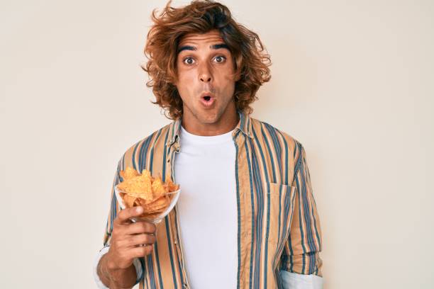 Young hispanic man holding nachos potato chips scared and amazed with open mouth for surprise, disbelief face Young hispanic man holding nachos potato chips scared and amazed with open mouth for surprise, disbelief face worried man funny stock pictures, royalty-free photos & images