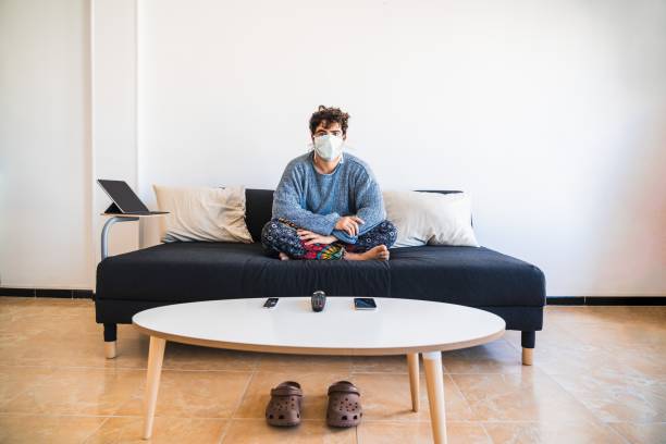 young hispanic man sits on the couch while protecting against covid-19 coronavirus due to it’s virulency and impact on media and society - pandemia doença imagens e fotografias de stock