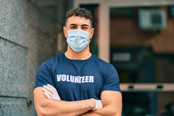 Young hispanic volunteer man with arms crossed wearing medical mask at the city. stock photo