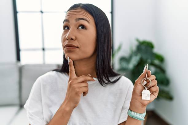 Young hispanic woman holding keys of new home thinking concentrated about doubt with finger on chin and looking up wondering stock photo