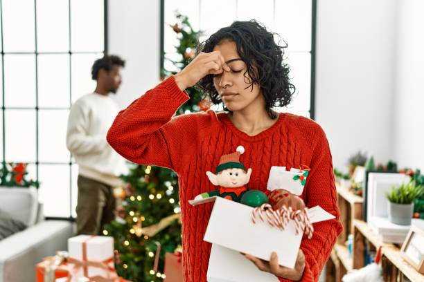 Young hispanic woman standing by christmas tree with decoration tired rubbing nose and eyes feeling fatigue and headache. stress and frustration concept. stock photo
