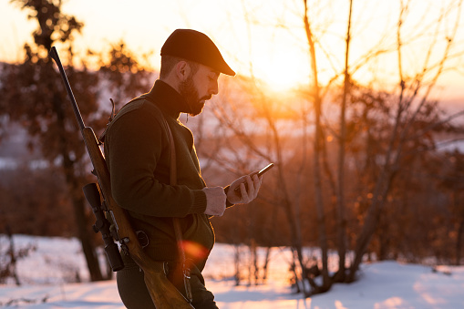 Young hunter is walking with smart phone in a field covered with snow and searching direction via GPS map during beautiful winter sunset.