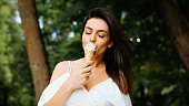 istock Young indian brunette woman in white summer dress eating vanilla ice cream in waffle cone, with closed eyes enjoying taste of food, outdoors. Beautiful elegant girl on summer vacation in park 1332339094