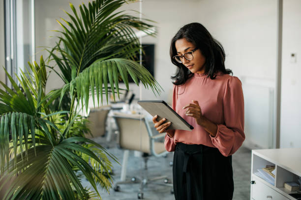 Young Indian Businesswoman Using Digital Tablet in Office Female executive with medium-length black hair wearing eyeglasses and businesswear standing outside modern meeting room and checking data on tablet. businesswoman stock pictures, royalty-free photos & images