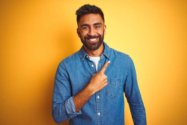 young indian man wearing denim shirt standing over isolated yellow background cheerful with a smile of face pointing with hand and finger up to the side with happy and natural expression on face - man pointing imagens e fotografias de stock