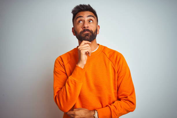 質問、ペンシブな表情を考えるあごに手を置いて、孤立した白い背景の上にオレンジ色のセーターを着た若いインド人男性。思慮深い顔で微笑む。疑わしい概念。 ストックフォト