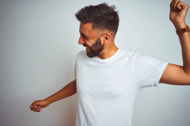 young indian man wearing t-shirt standing over isolated white background dancing happy and cheerful, smiling moving casual and confident listening to music - dancer white man on white imagens e fotografias de stock