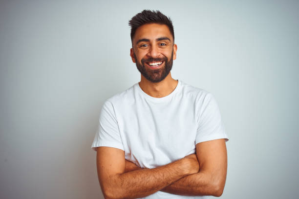 孤立した白い背景の上に立つTシャツを着た若いインド人男性は、カメラを見て腕を組んで微笑んでいる。ポジティブな人 ストックフォト